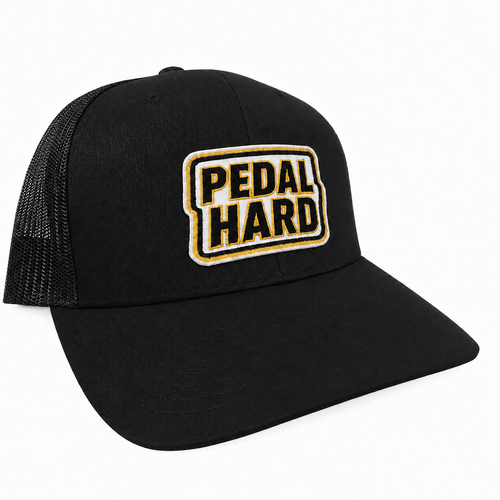 PEDAL HARD Flatlander black low-profile curved bill cycling trucker snapback with Athletic Yellow embroidered patch, front view.